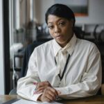 Serious African American businesswoman wearing formal white blouse and sitting at office desk with documents while looking at camera
