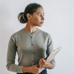 Serious ethnic female in formal wear with clipboard in hands standing against white wall and looking away in contemplation